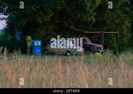 Alte russische Autos stehen am Rand eines Kornfeldes unter dem warmen Sommeruntergang. Neben ihnen ist der Tank. Stockfoto