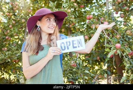 Eine glückliche Frau mit offenem Schild, um an einem sonnigen Tag auf einer nachhaltigen Obstplantfarm draußen für die Apfelernte zu werben. Fröhlicher Bauer zeigt auf Stockfoto