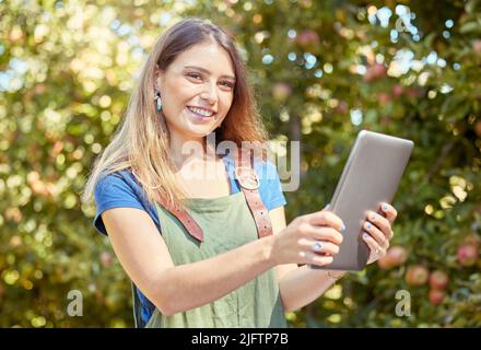 Eine Apfelbauerin, die steht und ein digitales Tablet benutzt, während sie ihre Pflanzen überprüft. Lächelnde Frau, die Technologie verwendet, um sich auf die Ernte auf ihrer Farm vorzubereiten Stockfoto