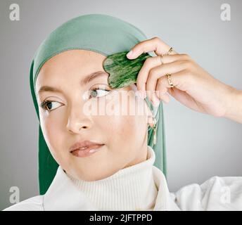 Ein Gua Sha, das auf einem jungen gesicht muslimischer Frauen verwendet wird, isoliert vor grauem Studiohintergrund. Frau mit einem Hijab oder Kopftuch, mit einem Anti-Aging Stockfoto