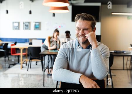 Lächelnder kaukasischer Geschäftsmann mit mittlerem Erwachsenenalter, der im kreativen Büro auf einem Stuhl sitzt Stockfoto