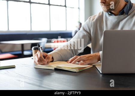 Mittelteil des kaukasischen Geschäftsmannes mit mittlerem Erwachsenenalter, der im Kreativbüro per Laptop Tagebuch schreibt Stockfoto