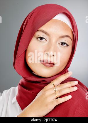 Porträt einer stilvollen muslimischen Frau mit Make-up und goldenen Ringen isoliert vor grauem Hintergrund. Freche junge Hijab-Dame, die sich in ihrem roten Schal sicher fühlt Stockfoto