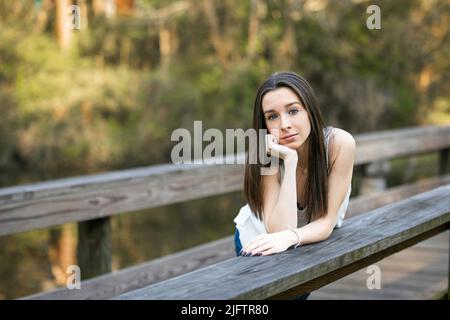 Eine große und dünne Brünette Teenager, die auf einer Parkbrücke steht und sich lehnt, wobei ihr Kopf mit ernstem Ausdruck auf ihren Händen ruht Stockfoto