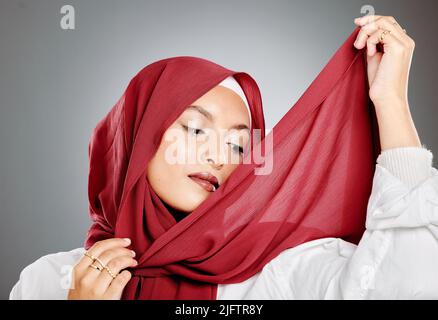 Eine elegante muslimische Frau, die einen roten Hijab oder ein Kopftuch trägt, das vor grauem Hintergrund mit Copyspace isoliert ist. Selbstbewusste Frau, die ihren Kopf in eine einhüllte Stockfoto