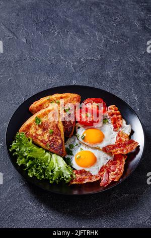 irisches Frühstück mit Spiegeleiern, Rascher- oder Speckscheiben, gebratenen Kartoffelfarn, gegrillten Tomaten und frischem Salat auf schwarzem Teller auf Betontisch, vertic Stockfoto