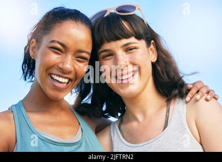 Porträt zweier lächelnder Freunde, die sich umarmen, während sie zusammen gegen den blauen Himmel stehen. Lächelnde, glückliche junge Frauen, die sich im Sommer im Freien anschmiegen und umarmen Stockfoto