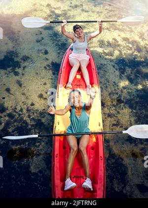 Oben Porträt von zwei verschiedenen jungen Frauen, die beim Kanufahren auf einem See jubeln und feiern. Begeisterte Freunde genießen Rudern und Kajakfahren auf einem Fluss Stockfoto