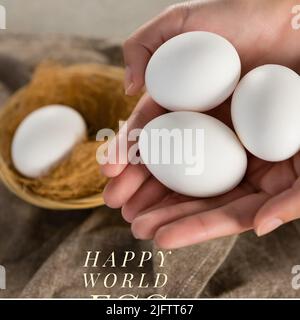 Beschnittene Hände einer kaukasischen Frau, die weiße rohe Eier hält, und Text zum Happy World Egg Day Stockfoto