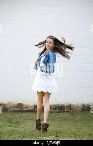 Ein Mädchen im Teenageralter mit Sommersprossen und langen, dunklen, strafferen Haaren, die draußen an einer weißen Wand herumlaufen und sich drehen Stockfoto