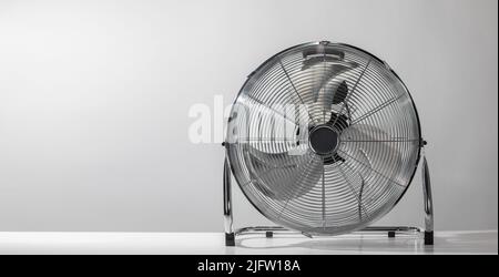 Silberner Metallbodenventilator auf dem weißen Boden zu Hause oder im Geschäft mit grauer Wand. Stockfoto