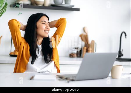 Glücklich entspannt positive junge chinesin, sitzt an einem Tisch in der Küche, kreuzt die Hände hinter dem Kopf, genießt die Pause während entfernter Online-Arbeit oder Studium, schaut beiseite, träumt, plant, lächelt Stockfoto