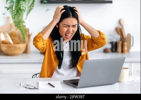 Frustriert traurig wütend asiatische junge Frau, Freiberufler, Manager, bekam ein negatives Ergebnis, gescheitert das Projekt, schaut verzweifelt auf Laptop-Bildschirm, hält ihre Hände auf den Kopf, haben die Situation gestresst, schreiend Stockfoto