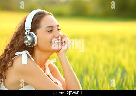 Fröhliche Frau, die Musik hört, mit kabellosen Kopfhörern, die auf einem Weizenfeld sitzen Stockfoto