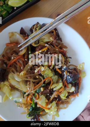 Japchae mit Karotten und Sesam auf dem weißen Teller auf dem Tisch.ist ein herzhaftes und leicht süßes Gericht aus gebratenen Glasnudeln und Gemüse. Stockfoto