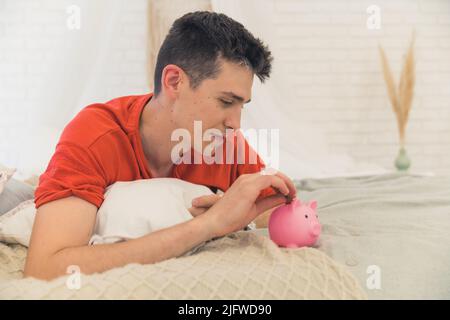 Der verantwortliche junge kaukasische Vater sorgte sich um die wirtschaftliche Situation, legte sich auf sein Bett und legte eine Münze in ein rosafarbenes Sparschwein. Symbolische Aufnahme. Hochwertige Fotos Stockfoto