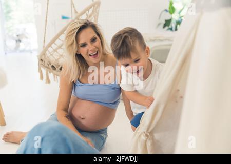 Fröhliche, schwanger Mutter und ihr Sohn sitzen auf dem Boden und haben Spaß. Hochwertige Fotos Stockfoto