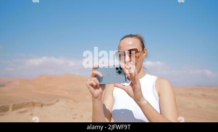 Touristenfrau, die in der Wüste steht und das Handy zum Schießen benutzt. Aktion. Junge Frau, die auf ihrem iPhone ein Bild von der Wüste gemacht hat Stockfoto