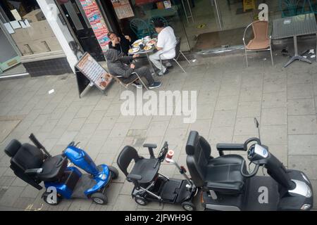 Aus der Luft gesehen, essen drei Personen an einem Tisch vor einem Café zusammen mit drei Mobilitäts-Scootern auf der Walworth Road im Süden Londons, am 5.. Juli 2022, in London, England. Stockfoto