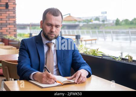 Ein fokussierter Geschäftsmann im Alter von 40s Jahren schreibt Notizen in ein persönliches Tagebuch, plant einen Arbeitstag, prüft einen Zeitplan oder schreibt wichtige Informationen von Hand Stockfoto