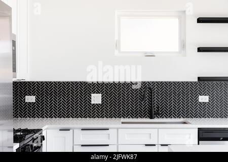 Ein Küchenwaschbecken Detail mit einem schwarzen Wasserhahn, schwarzem Marmor Fischgrätenhinterspritzer und einer weißen Granit-Arbeitsplatte aufgenommen. Stockfoto