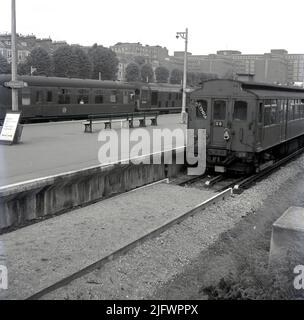 1960s, historische Ansicht von Kensington Olympia Bahnhof und U-Bahnstation, Kensington, London, England. VEREINIGTES KÖNIGREICH. Ein Schild und ein Pfeil auf dem Bahnsteig sagen, dass sich Züge nach Earls Court und High Street Kensington auf der rechten Seite befinden, wo ein oberirdisch anfahrender U-Bahn-Zug wartet, mit der Welt Olympia im Vorderfenster. Ein Hauptzug wartet auf dem gegenüberliegenden Bahnsteig, Nr. M 34544 und Wagen Nr. D 5019. Stockfoto