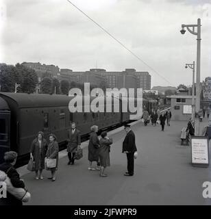 1960s, historisch, Passagiere auf dem Bahnsteig am Kensington Olympia Zug- und U-Bahnhof, Kensington, London, England. Das Vereinigte Königreich hat einen Hauptzug gestartet. Zwei Passagiere sprachen mit einem uniformierten Bahnhofsmanager auf dem Bahnsteig. In weiter Ferne ein Courage Pub, The Hand & Flower an der Straßenbrücke. Stockfoto