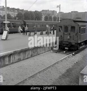 1960s, historische Ansicht von Kensington Olympia Bahnhof und U-Bahnstation, Kensington, London, England. VEREINIGTES KÖNIGREICH. Ein Hauptzug wartet auf dem Bahnsteig, während ein Schild sagt, dass Züge nach Earls Court und High Street Kensington auf der rechten Seite sind, wo eine oberirdische U-Bahn wartet, mit der Welt Olympia im Fenster. Stockfoto