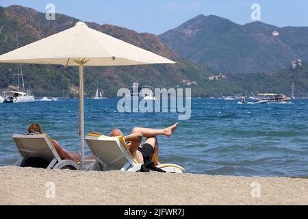 Paar bräunen am Strand im Sommerresort. Mann und Frau liegen auf Liegestühlen unter Sonnenschirmen auf Meereswellen und im Hintergrund der Bergküste Stockfoto