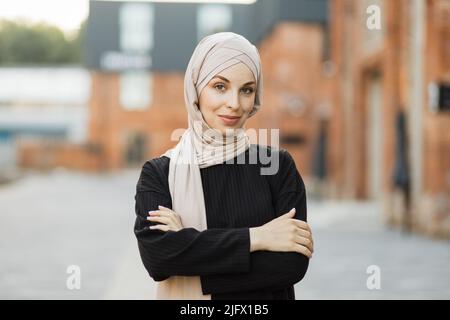 Porträt einer jungen muslimischen Frau, die in der Stadt einen Hijab-Kopftuch trägt, während sie die Kamera anschaut. Nahaufnahme der fröhlichen Frau, bedeckt mit einem Kopftuch, das im Freien lächelt. Casual islamischen Mädchen in der Stadt. Stockfoto