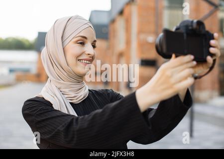 Junge muslimische Bloggerin in trendigen Kleidern und Hijab geht auf der Stadtstraße, nimmt sich selbst auf Video auf und hält Kamera in der Hand. Arabisches Mädchen macht Bilder von sich selbst vor der Kamera. Lifestyle Selfie-Hintergrundbeleuchtung Stockfoto