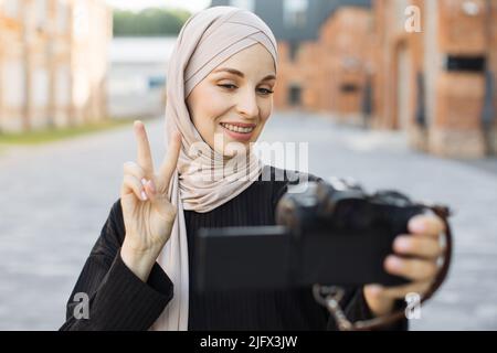 Junge muslimische Frau, die mit einer Videokamera eine Reise mit einem Lächeln aufzeichnet. Glücklich und aufgeregt 20s arabische Mädchen Reisenden tragen schwarze Kleidung mit Hijab, winken zu Internet-Publikum Zeichen des Friedens zeigen Stockfoto