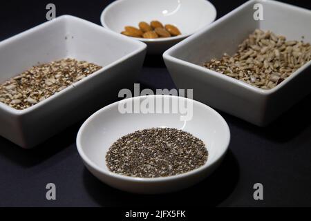 Mandeln, Chia und andere gesunde vegane Samen in weißen Porzellanschalen auf schwarzem Hintergrund Stockfoto