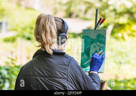 Barcelona, Spanien - 22. April 2022: Künstlerin malt ein Bild im botanischen Garten von Barcelona (Spanien), selektiver Fokus auf den Kopfhörer. Stockfoto