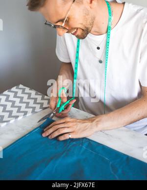 Ein lächelnder Schneider schneidet Stoff in einem Heimstudio. Ein autodidaktischer Seemann in weißem T-Shirt und Brille arbeitet mit Stoff, Muster und Kreide. Stoffschnitt Stockfoto