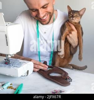 Nähen von Kleidung für Haustiere. Schneiderbekleidung für Katzen. Ein lachender Schneider in weißem T-Shirt näht Kleidung für seine abessinier-Katze. DIY-Geschäftskonzept Stockfoto