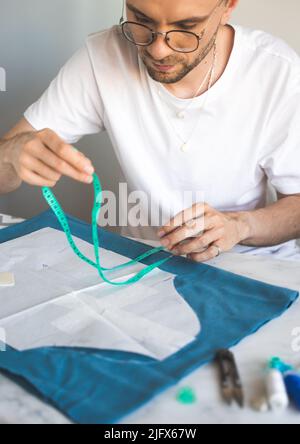 Ein autodidaktischer Seemann in weißem T-Shirt und Brille arbeitet mit Stoff, Muster und Kreide. Ein Schneider verwendet bei seiner Arbeit zu Hause ein Maßband Stockfoto