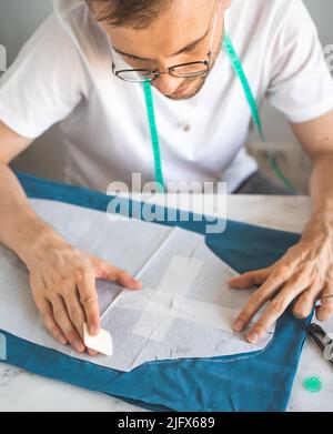 Ein autodidaktischer Seemann in weißem T-Shirt und Brille arbeitet mit Stoff, Muster und Kreide. Ein Schneider überträgt eine Vorlage in ein Heimstudio auf Stoff. Stockfoto