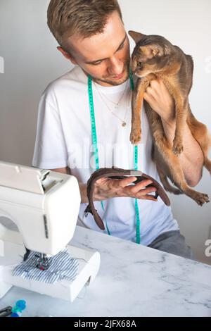 Schneiderbekleidung für Katzen. Nähen von Kleidung für Haustiere. Ein lächelnder Mann in weißem T-Shirt näht Kleidung für seine abessinier-Katze. Konzept für kleine Unternehmen Stockfoto