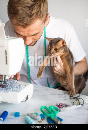 Nähen von Kleidung für Haustiere. Schneiderbekleidung für Katzen. Ein lächelnder Mann näht Kleidung für seine abessinier-Katze. Das Konzept eines kleinen und DIY-Unternehmens. Stockfoto