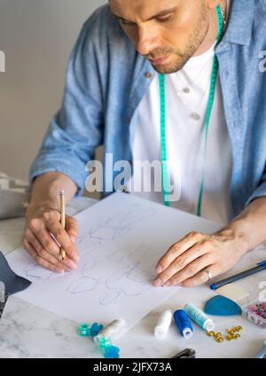 Ein Mann in einem blauen Hemd macht eine Skizze für das Nähen von Kleidung als Hobby. Ein autodidaktischer Seemann mit einem Maßband am Hals arbeitet am Tisch. Stockfoto