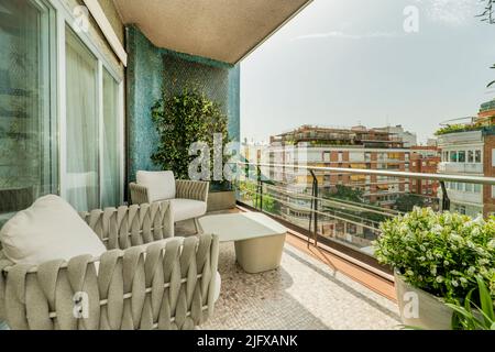 Terrasse mit grauen Stoffsesseln, Metallgeländer, Pflanzgefäß, vertikalem Garten an einer blauen Wand und Blick auf die Stadt Stockfoto