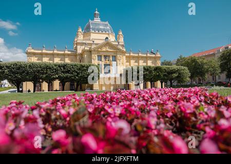 Schönes Gebäude der Kunstgalerie im Zentrum von zagreb am Kralj Tomislav Park an einem heißen Sommertag. Gelbes Gebäude mit roten Blumen im Vordergrund. Stockfoto