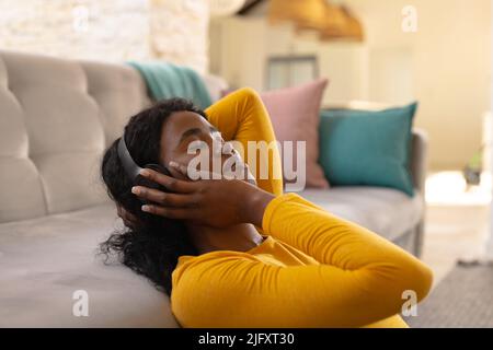 afroamerikanische junge Frau mit geschlossenen Augen, die Musik auf dem Kopfhörer genießt, während sie sich auf das Sofa lehnt Stockfoto