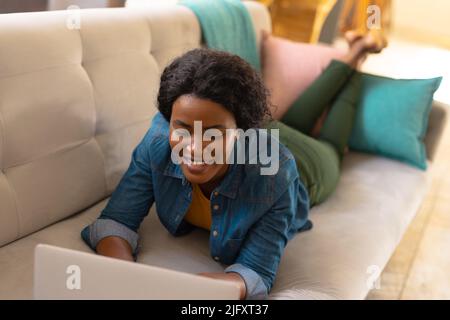 Lächelnde junge afroamerikanische Frau mit Laptop, während sie zu Hause auf dem Sofa liegt, Platz kopieren Stockfoto