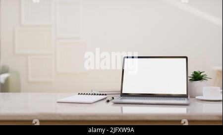 Arbeitsbereich mit Laptop-Computer, leerem Bildschirm, spiralförmigem Notizblock, Zubehör und Kopierplatz auf Marmor-Tischplatte über unscharfem, hellem Wohnzimmer in b Stockfoto