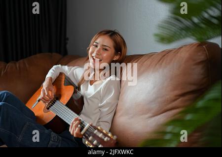 Attraktive und talentierte Millennial Asian Mädchen sitzen auf bequemen Sofa im Wohnzimmer, singen und spielen ihre klassische Gitarre. Kreative Aktivität Konz Stockfoto