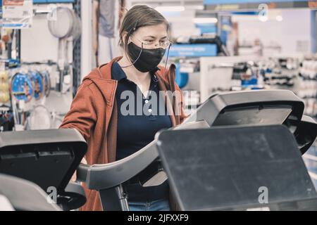 Seriöse Frau in Brille und Maske gegen Virus kauft große Laufband in einem Geschäft Stockfoto