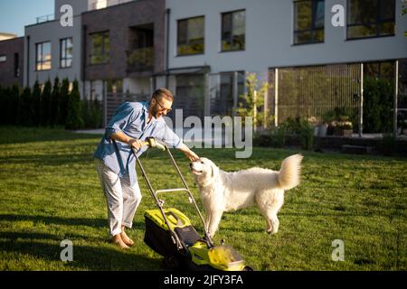 Der Mann spielt mit einem Hund, während er den Rasen im Hinterhof bewegt Stockfoto
