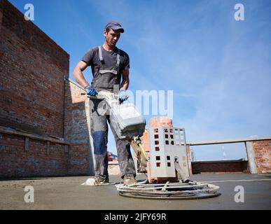 Die gesamte Länge des Arbeiters mit einer Kelle, während der Fußboden im neuen Gebäude unter blauem Himmel Estriche. Männlicher Arbeiter, der auf der Baustelle eine Betonoberfläche mit einer Bodentrichschleifmaschine fertig stellte. Stockfoto